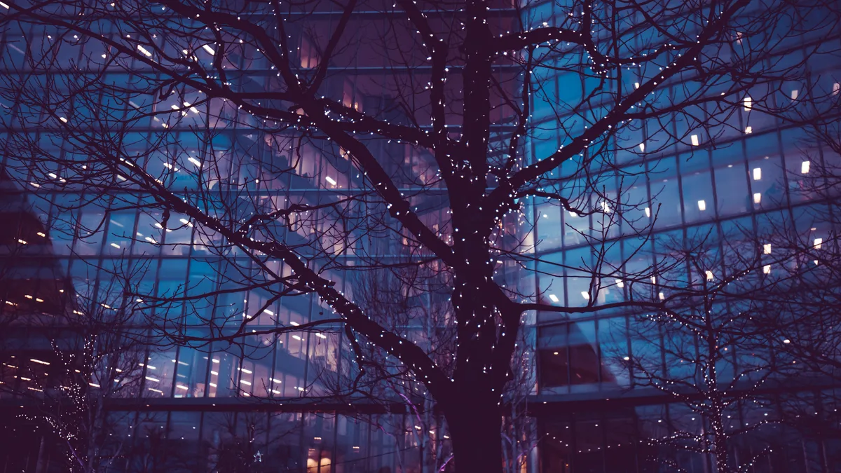 Trees wrapped in string lights in front of a glass office building at night