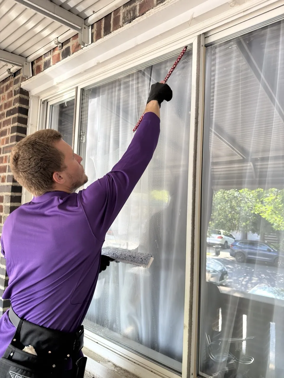 Purple Window Cleaning technician in uniform washing a residential window with a squeegee and washer